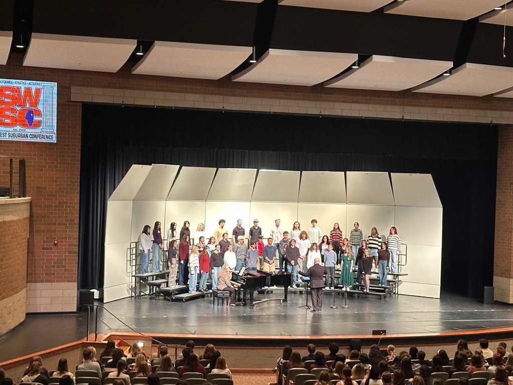 Choir on stage at school auditorium with audience seated in front. Stage has piano, microphone and sign.