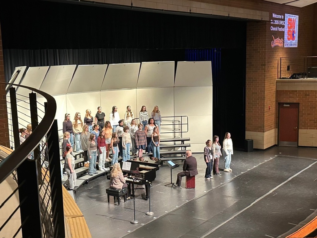 Choir performing on stage with pianist in front of audience. Two large banners hanging in the background.