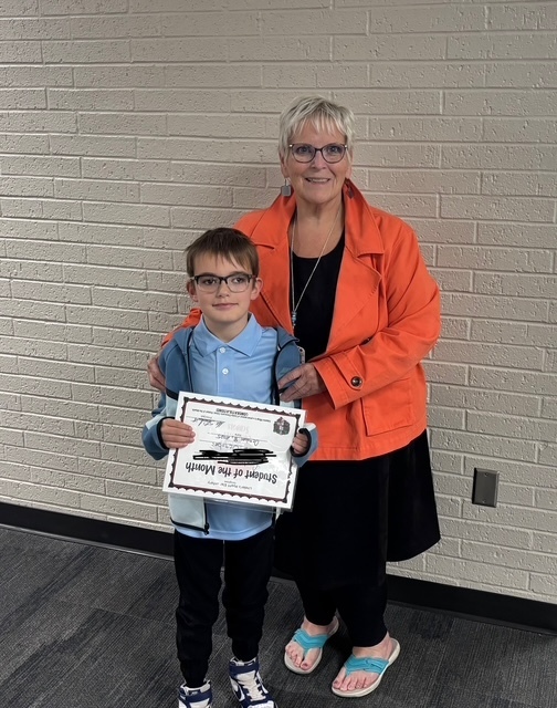 Female teacher with orange jacket standing next to a male student with glasses and a blue shirt.. Both are smiling. The student is holding a student of the month certificate.