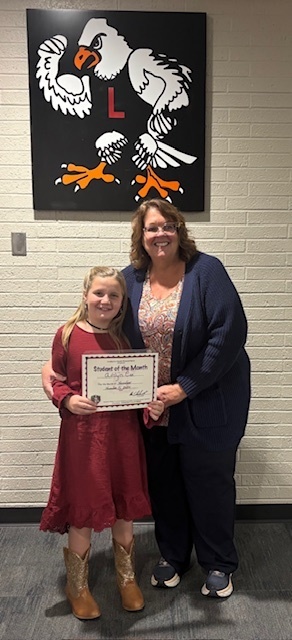 Female teacher with brown hair and a blue sweater standing next to her third grade student.  The third grade student is holding her student of the month certificate.