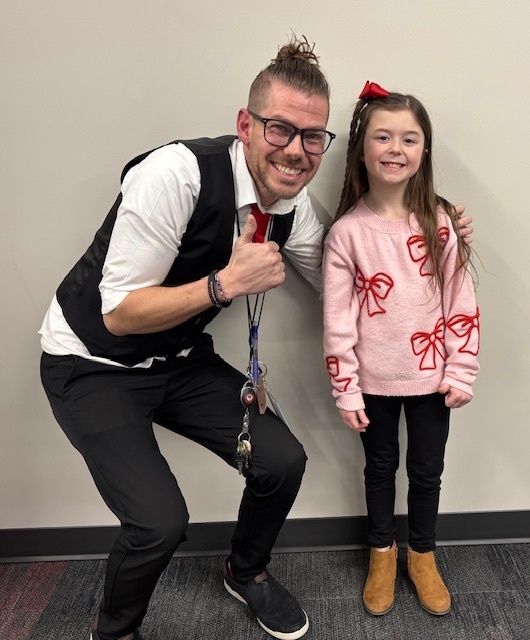 A male teacher wearing glasses, black pants, white shirt, black vest is smiling with his thumb raised as he leans down next to his female second grader. She has brown hair, is smiling, and is wearing a pink sweater with ribbons, dark pants and brown boots. She just received an award for student of the month.