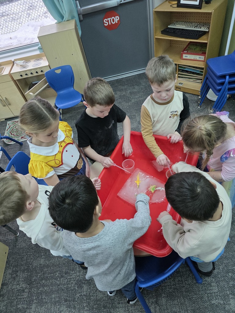 A group of preschool students gathered around a red sensory table, using pipets to put warm water on a block of  ice with plastic ocean animals frozen in  it.