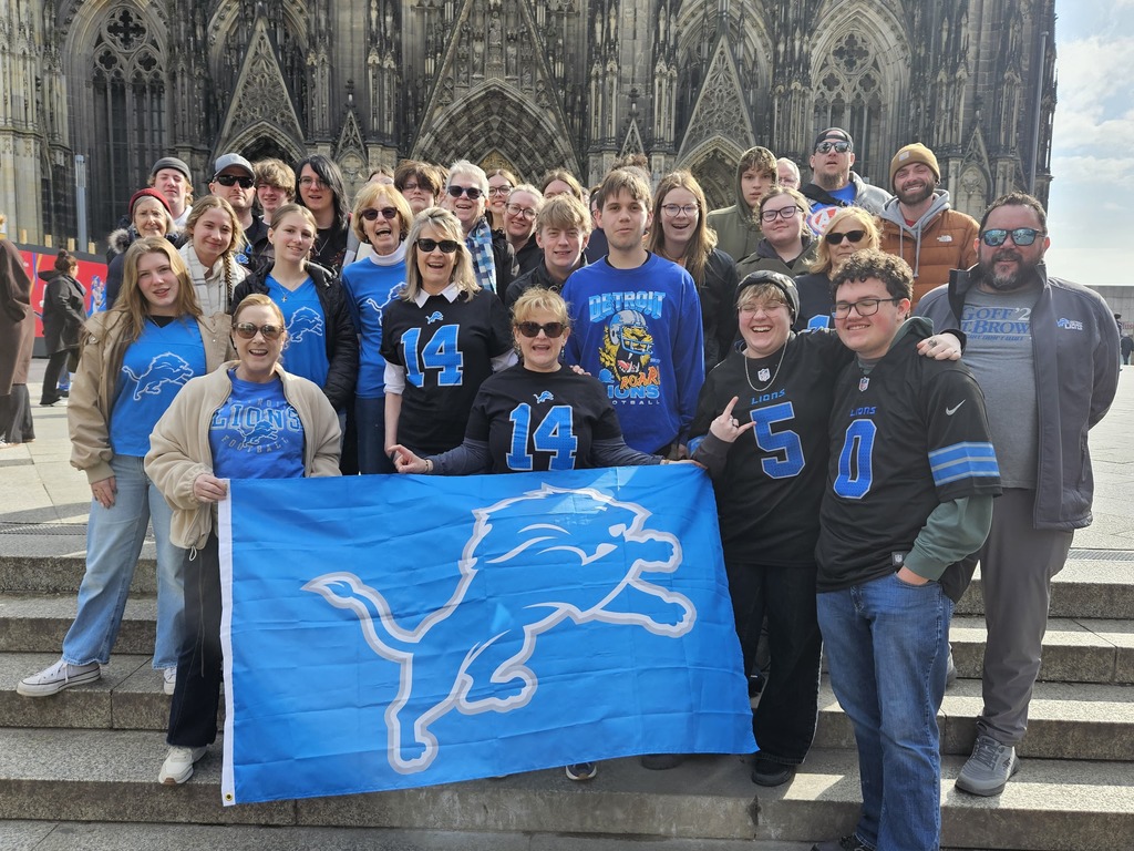 Group of students standing in front of Cologne Cathedral in Lions shirts and holding a Detroit Lions flag.