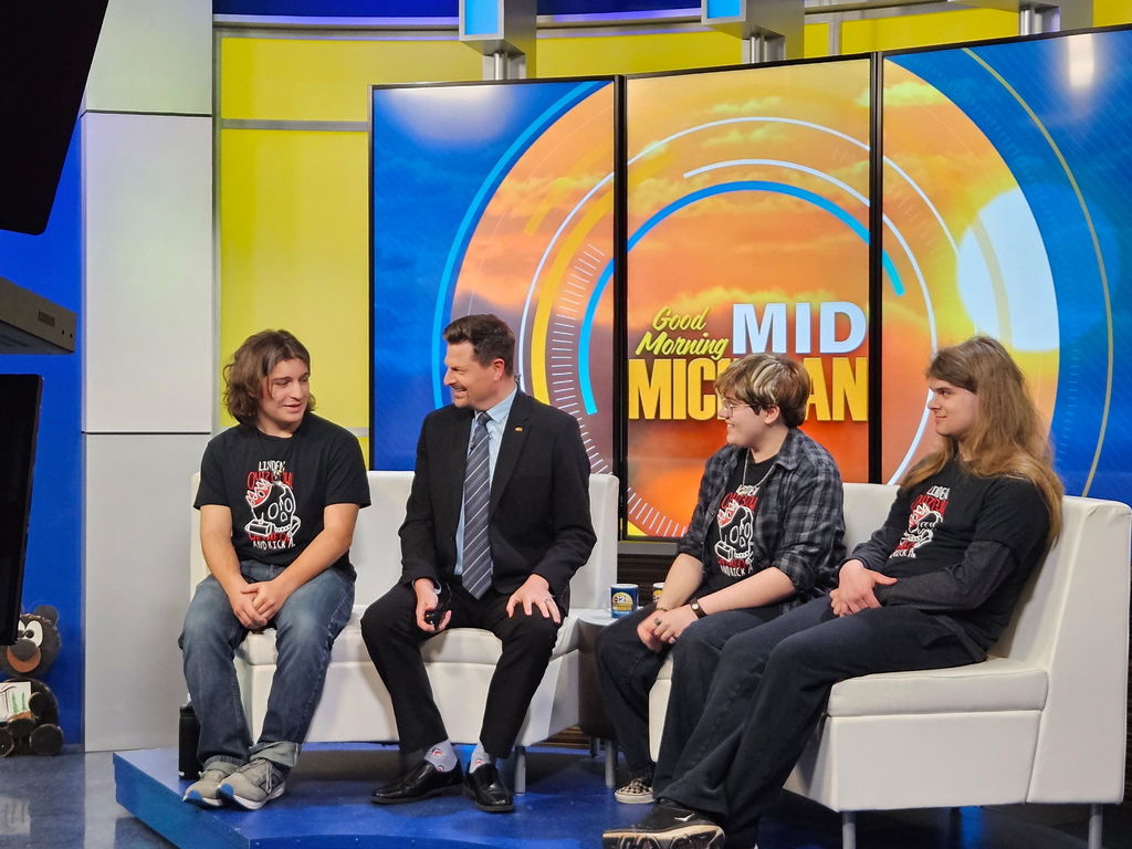 Three students wearing Linden Quiz Bowl with a news anchor from Channel 12 sitting on the morning news set