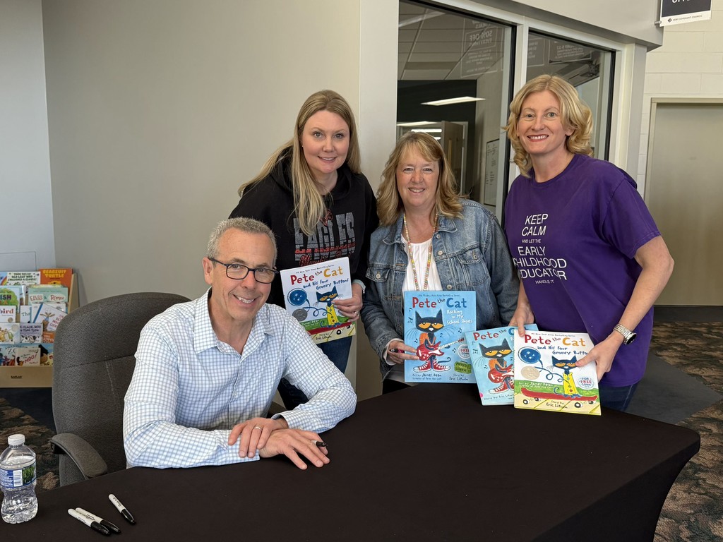An author/artist sits at a black table, smiling alongside three women who are holding up several "Pete the Cat" children's books. One woman wears a purple t-shirt that reads, "Keep Calm and let the Early Childhood Educator handle it." The group is in a brightly lit indoor space, possibly a school or library, with bookshelves visible in the background.