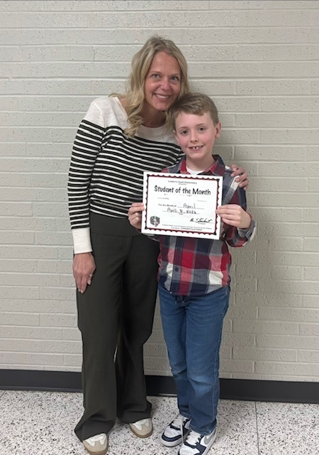 Female teacher wearing a long sleeved white sweater with black horizontal lines is smiling, standing with her hand on her student's shoulder.  The student is holding a student of the month certificate and  wearing a black and red checkered shirt.  