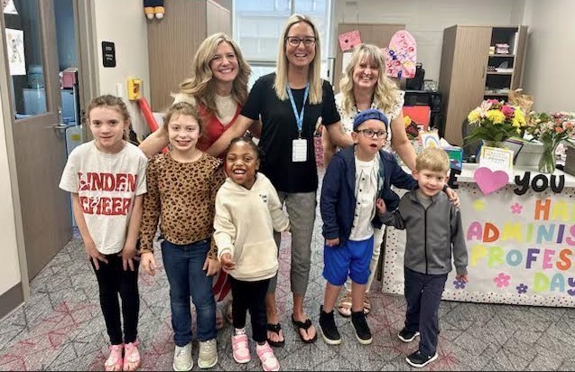 Three adults are standing behind five children.  All the people are smiling.  They are celebrating administrative assistant's day.