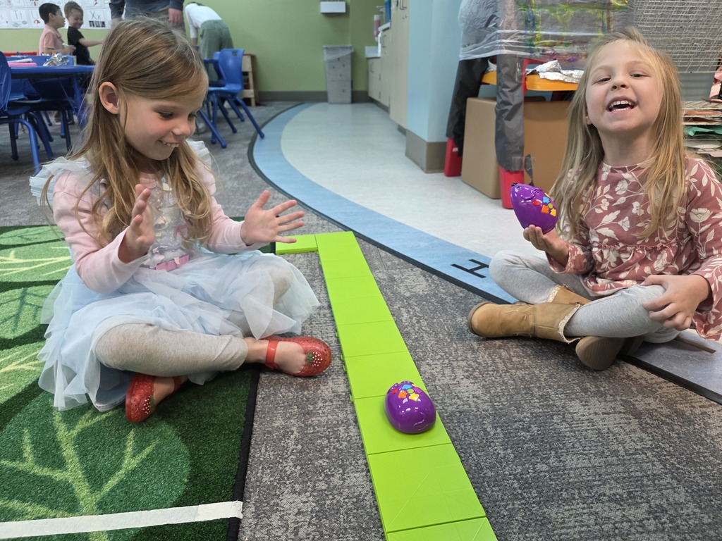 Two smiling students sit on either side of a connected line of green squares.  A programable coding mouse is in the middle of the line.  