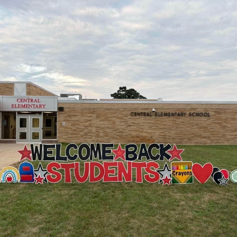 Welcome back students colorful sign with box of crayon and blue backpack in front of school building.