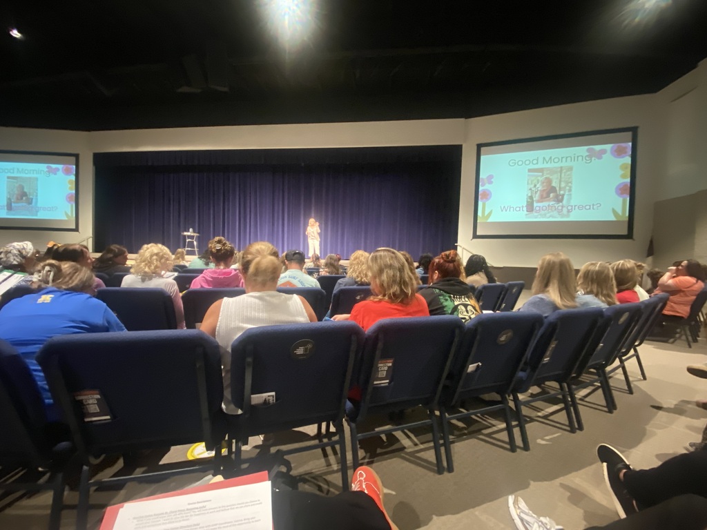 Audience members sit in rows of blue padded chairs inside an auditorium, looking toward a speaker standing on stage. Large projection screens flanking the stage display a presentation slide titled "Good Morning" with the text "What's going great?"