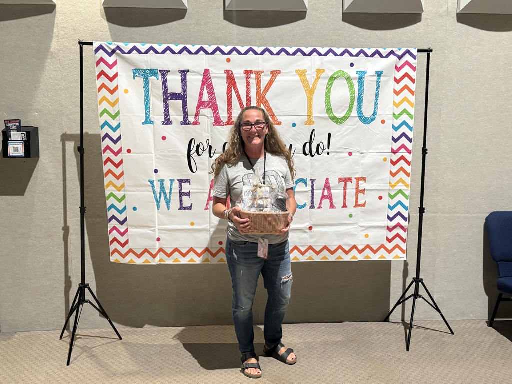 A smiling woman wearing glasses, a grey t-shirt, and jeans holds a wrapped gift basket while standing in front of a colorful backdrop. The backdrop features bright lettering that reads, "Thank You for all you do! We appreciate..." surrounded by a colorful chevron border.