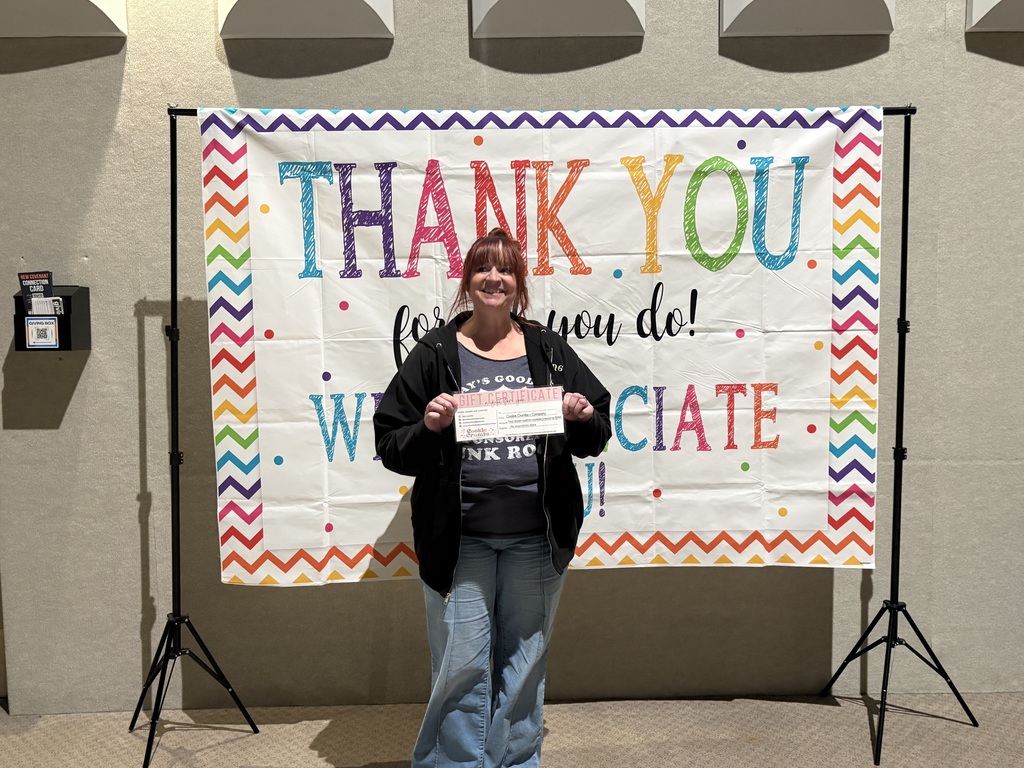 A smiling woman with reddish-brown hair, wearing a black zip-up hoodie and jeans, holds up a pink gift certificate in front of a colorful backdrop that reads, "Thank You for all you do! We appreciate you!".