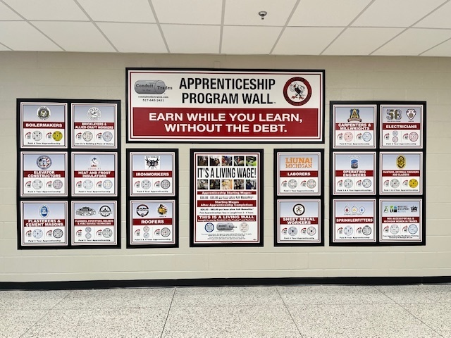 A school hallway wall featuring an "Apprenticeship Program Wall" bulletin board display with the header "Earn while you learn, without the debt," surrounded by numerous informational posters about various career and apprenticeship opportunities.