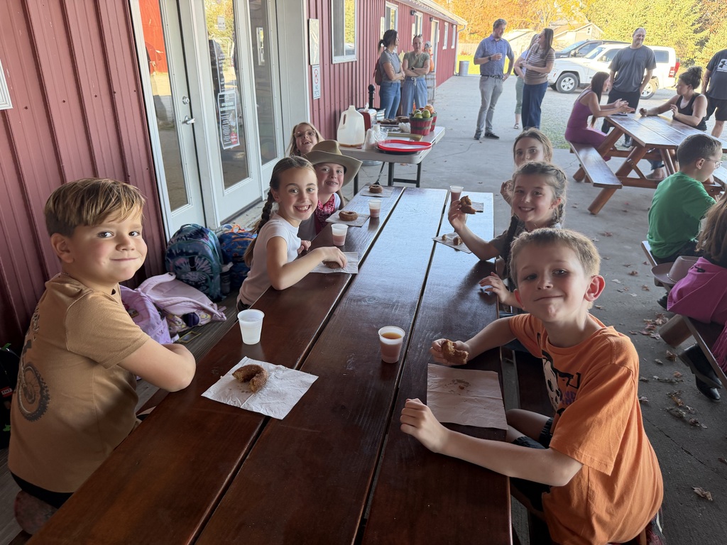 Elementary students smile while seated around a wooden picnic table eating cider donuts outdoors next to a red farm building.