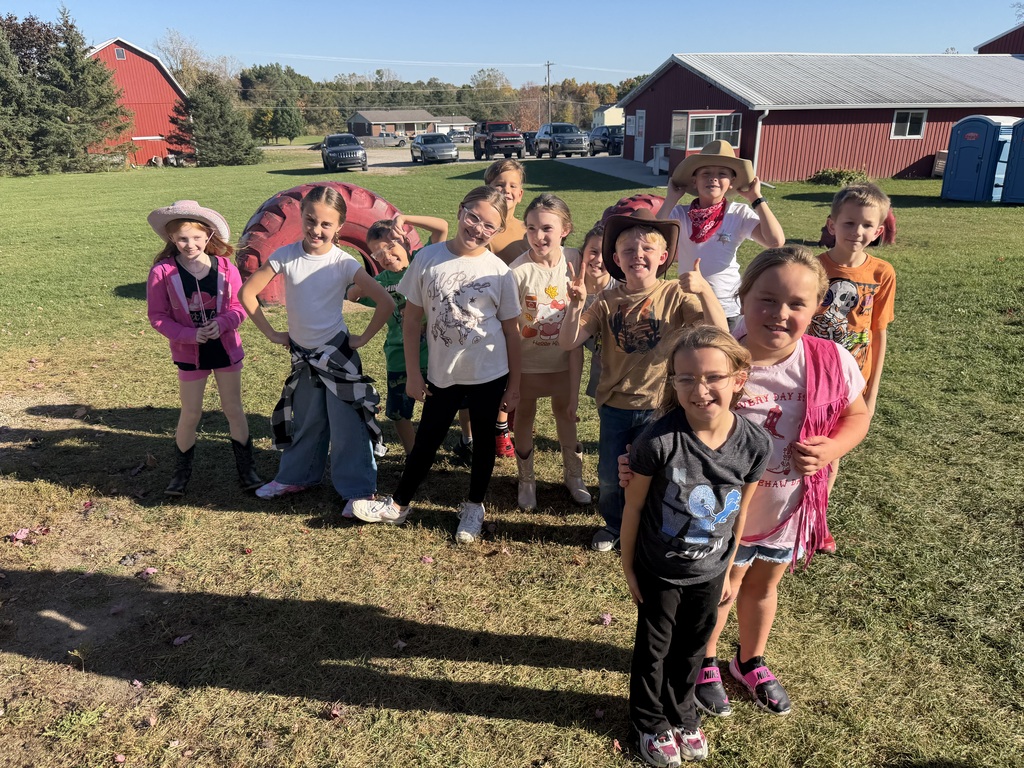 Elementary students dressed in Western hats and boots smile and pose together outdoors on a grassy lawn in front of a red barn.