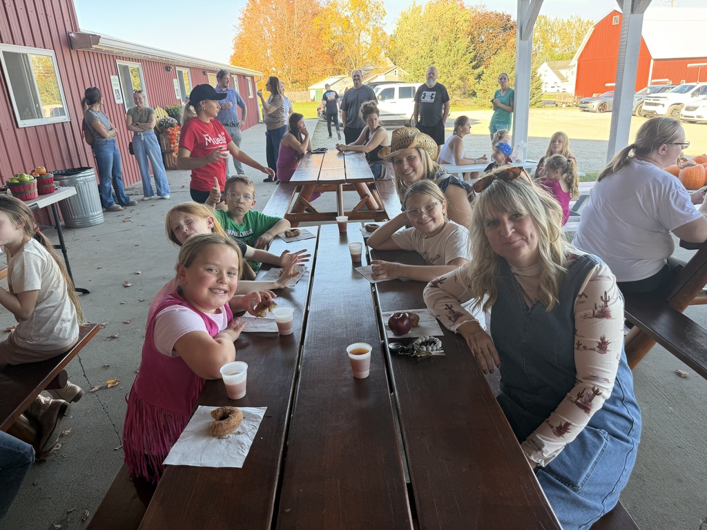 Students and chaperones sit together at long wooden picnic tables under a covered patio, enjoying cider and donuts during a field trip.
