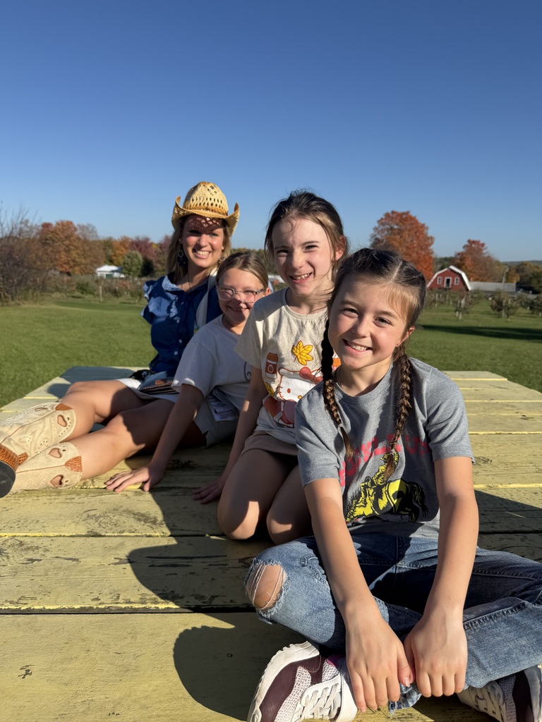 A teacher wearing a straw cowboy hat sits on a yellow wooden table outdoors alongside three smiling elementary students.