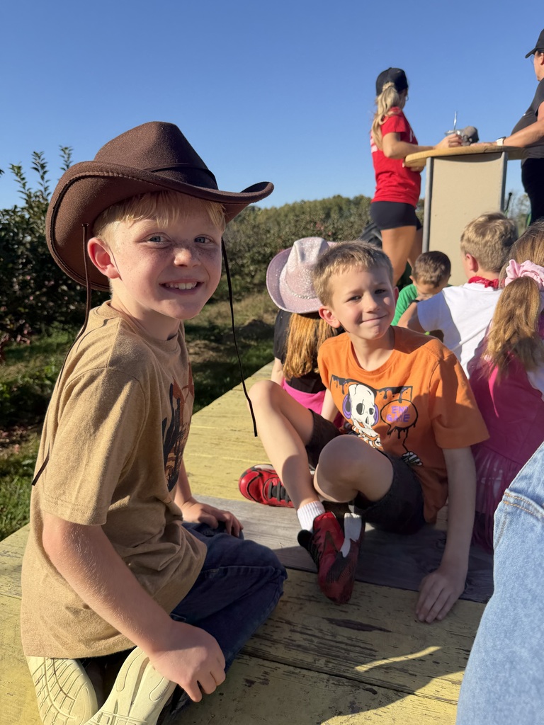 Two young boys smile while sitting on an outdoor wooden table, with one wearing a brown cowboy hat and the other an orange Halloween shirt.