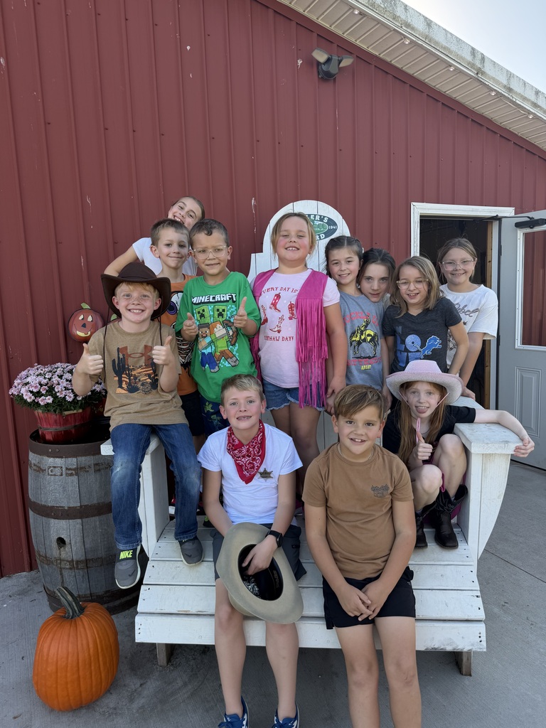 A group of elementary students dressed in cowboy hats and Western outfits pose on and around an oversized white Adirondack chair next to a red barn wall decorated with pumpkins.