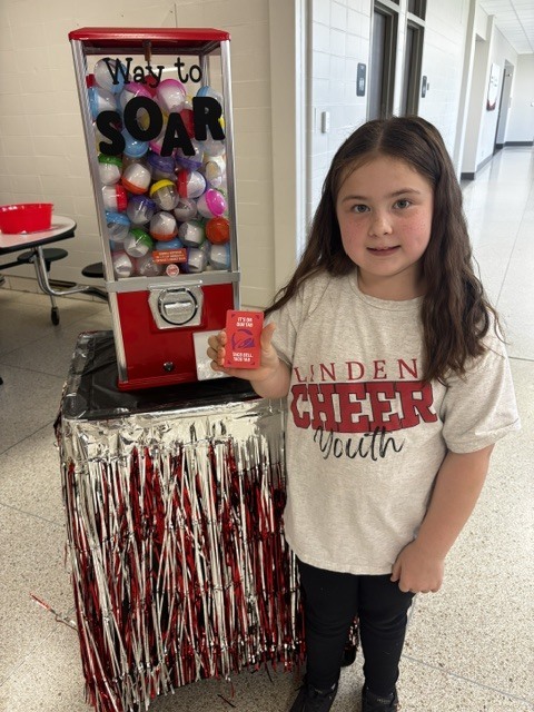 A student wearing a Linden Cheer Youth t-shirt holding up a red reward ticket in front of a Way to SOAR prize machine.
