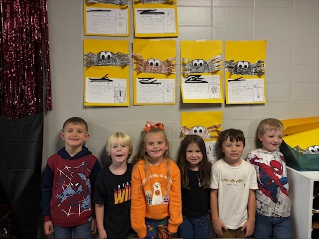 Six elementary students standing in a row in front of a classroom wall displaying spider craft projects on yellow paper.