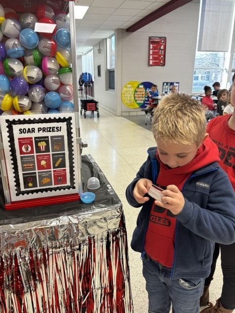 An elementary student opening a small prize slip next to a prize capsule machine labeled SOAR Prizes