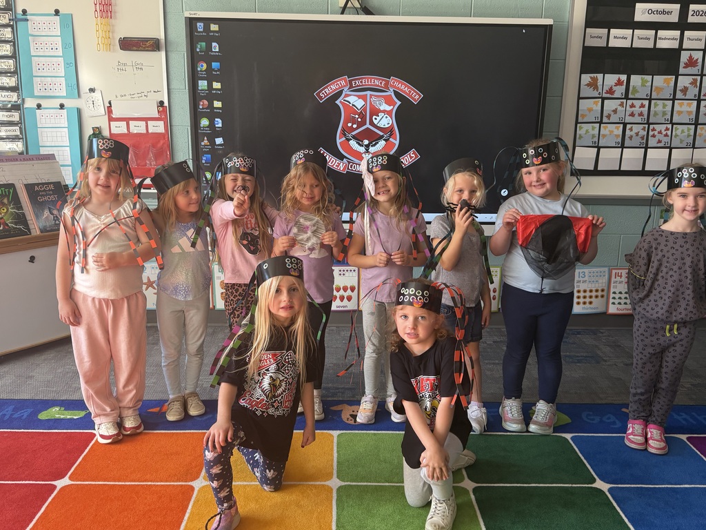 Group of elementary school girls wearing paper spider headbands standing in front of a classroom screen displaying the Linden Community Schools logo.