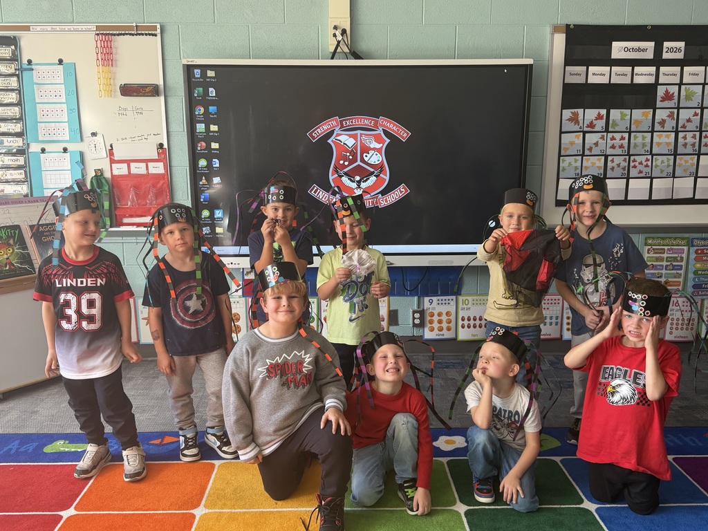 Group of elementary school boys wearing paper spider headbands posing in front of a classroom screen displaying the Linden Community Schools logo.