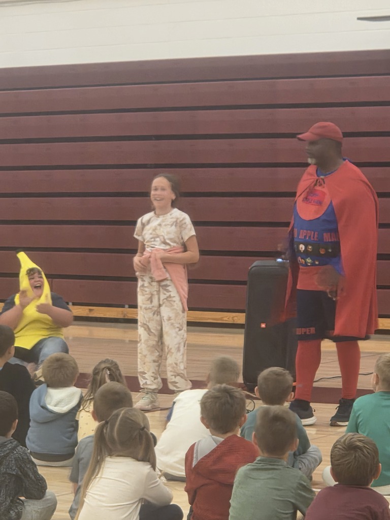 Elementary students sit on the gym floor facing the front of the gym during a school assembly featuring Super Apple Man. Students gather to learn about the importance of eating fruits and vegetables.