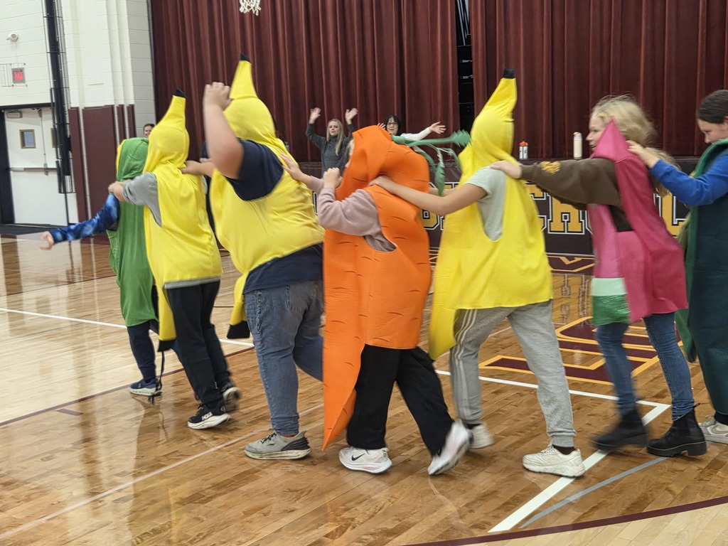 A group of elementary students pose together in the gym, including students wearing colorful fruit and vegetable costumes, as part of the school's Super Apple Man visit.