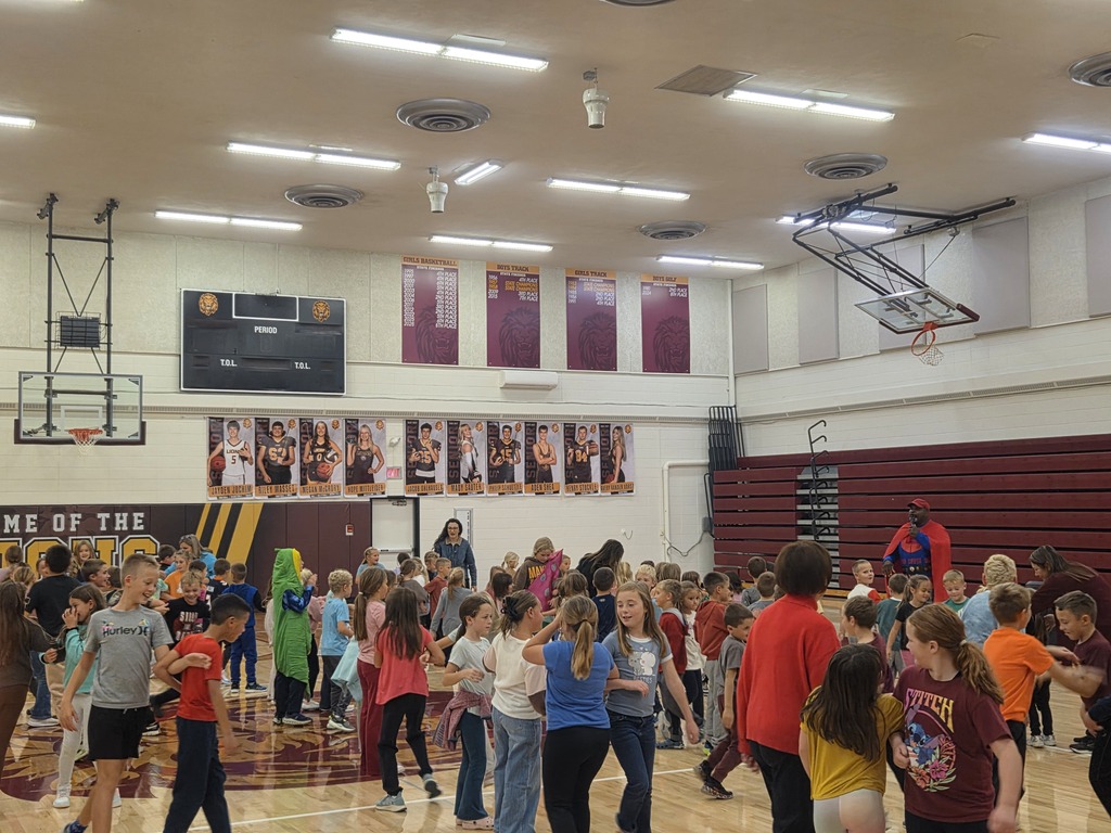 A group of elementary students pose together in the gym, including students wearing colorful fruit and vegetable costumes, as part of the school's Super Apple Man visit.