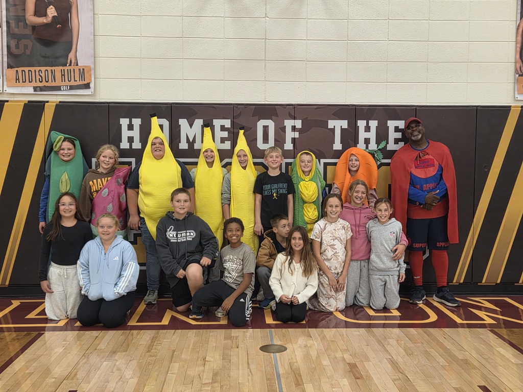 Elementary students pose in the school gym wearing colorful fruit and vegetable costumes, including banana, carrot, and other produce-themed outfits. Super Apple Man stands at the right of the group.