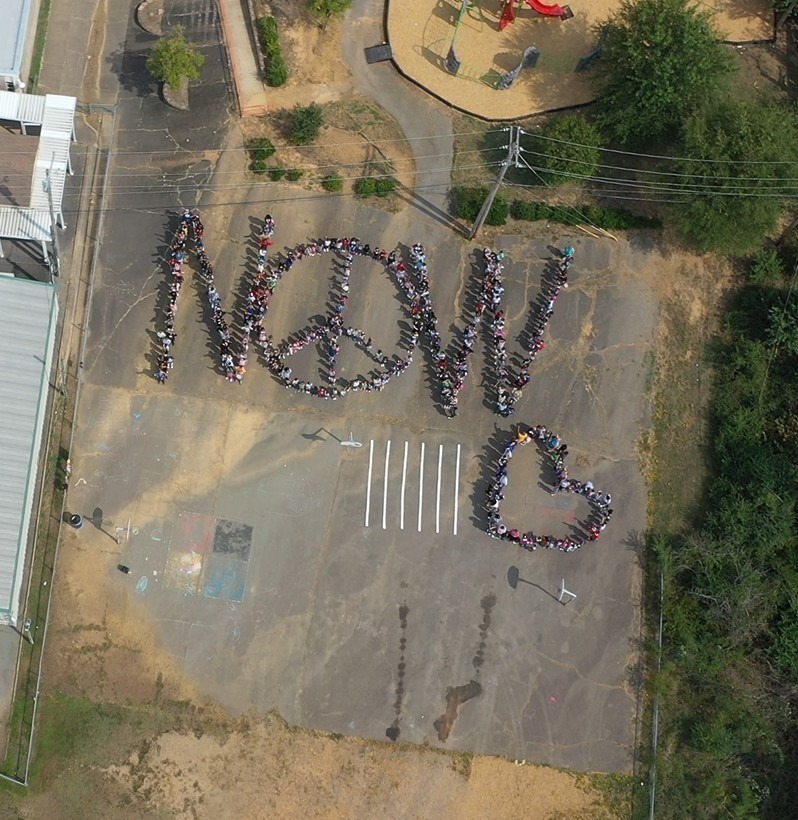 Students and staff at McDermott Elementary gather to create an aerial image of Now with a peace sign and a heart