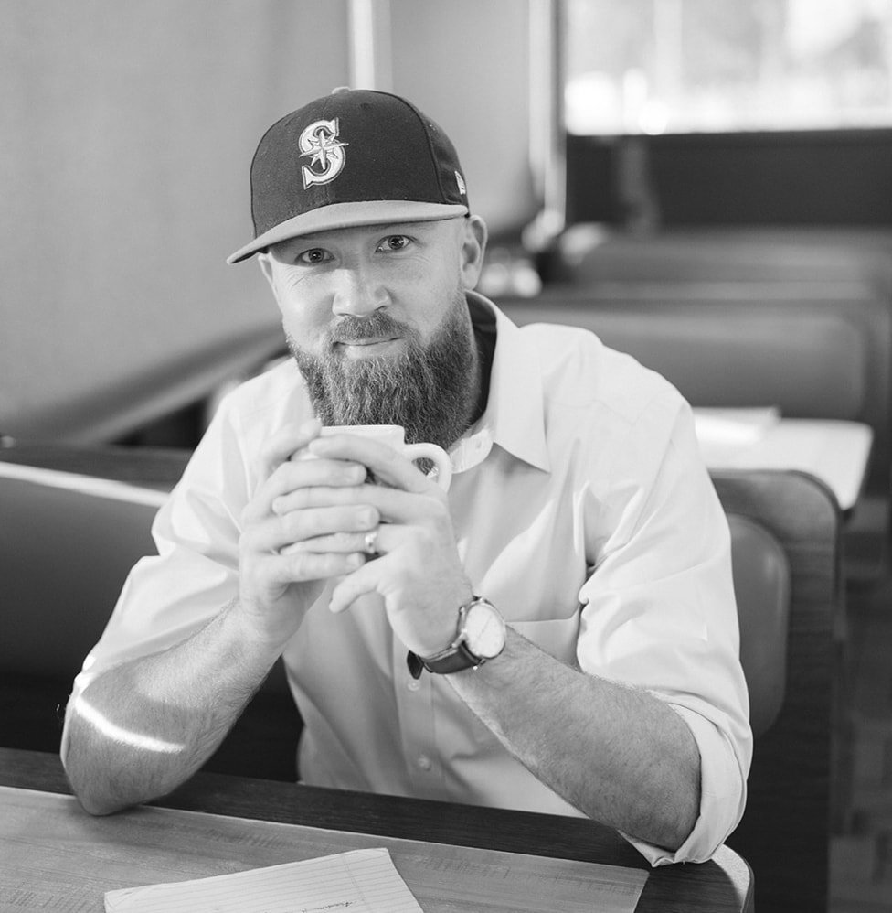 A black and white headshot of author Eli Cranor.  He is sitting in a dinner with a Seattle Mariners cap on holding a coffee cup.