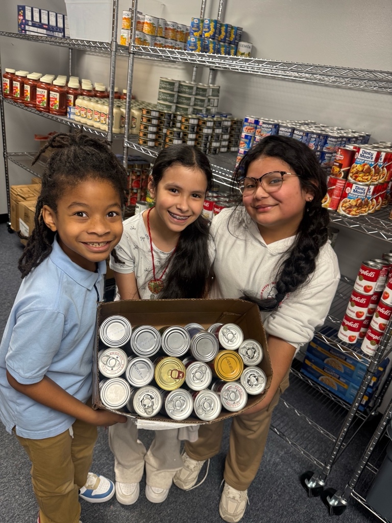 Fourth grade students stand together in front of shelves in the Chicot Elementary food pantry. They are smiling and posing after helping stock donated items. The shelves behind them are filled with food and household supplies.