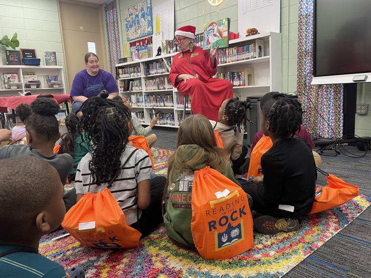 kids sitting at the carpet listening to the librarian read with their back packs full of books
