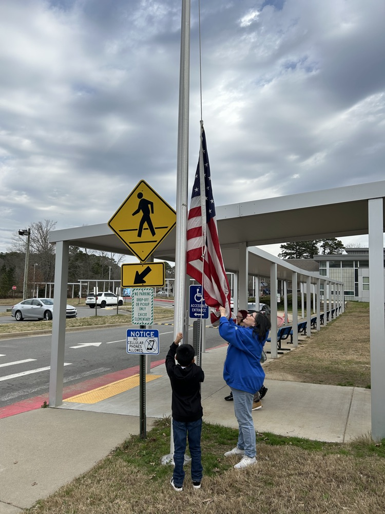 An adult and a student raise the American flag together in front of the school.l