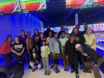 A large group of people is gathered at a bowling alley during what looks like a fun group outing or celebration. The group is posing together in front of the bowling lanes under colorful blue and purple neon lighting. Several bowling score screens are visible overhead, and many people are smiling at the camera. Some are standing while a few kneel in the front row, creating a casual and cheerful group photo atmosphere. Everyone is dressed comfortably, with a mix of casual outfits and bowling shoes visible.