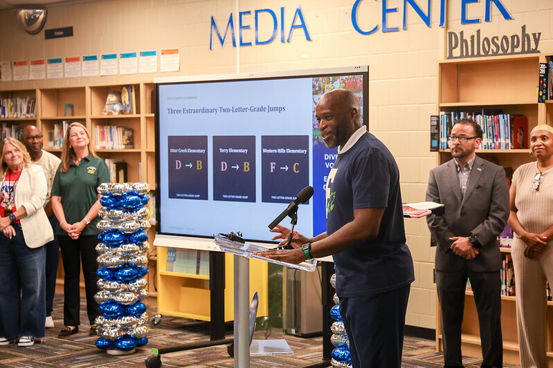 Photo of Dr. Wright at podium with staff and board members standing nearby; monitor that shows grades progressing from d to b and f to c