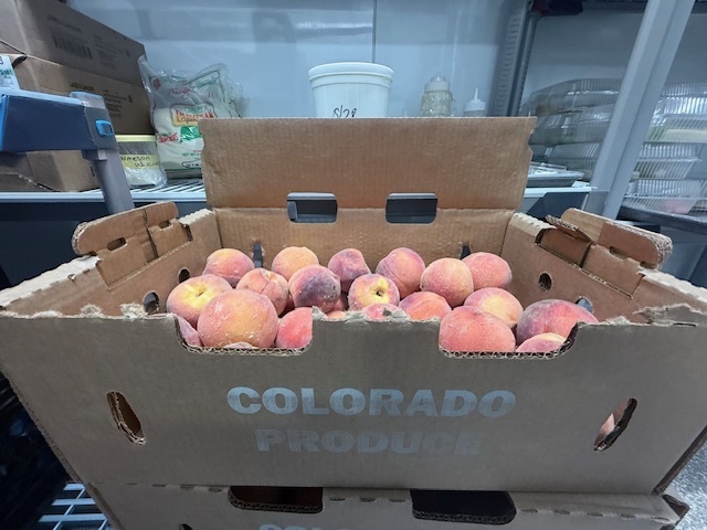 Fresh, fuzzy pink and yellow peaches fill a cardboard box labeled "Colorado Produce" in a kitchen workspace.