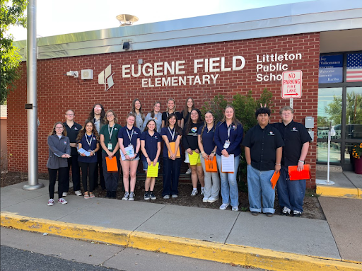 A group of approximately 16 students and adults are posing in front of a brick building with a sign that reads EUGENE FIELD ELEMENTARY and Littleton Public Schools.