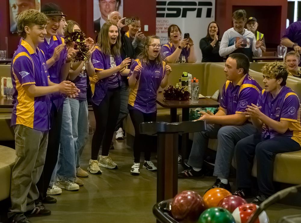A team of high school bowlers in purple and gold jerseys cheers for a teammate at a bowling alley.