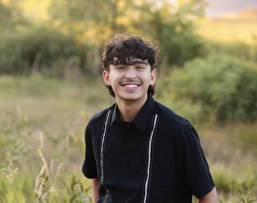 A smiling young man with curly dark hair and a mustache wearing a black button-up shirt in an outdoor grassy field.