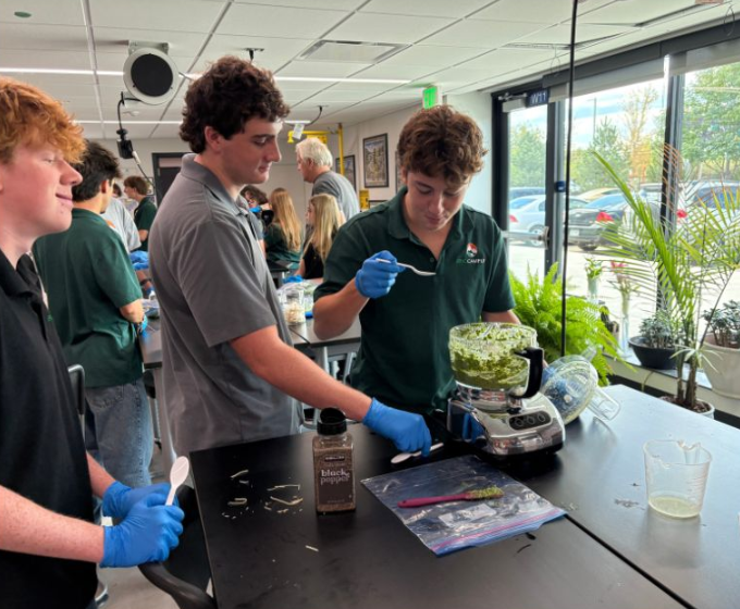 Students wearing blue gloves taste-test freshly blended pesto from a food processor in a classroom.
