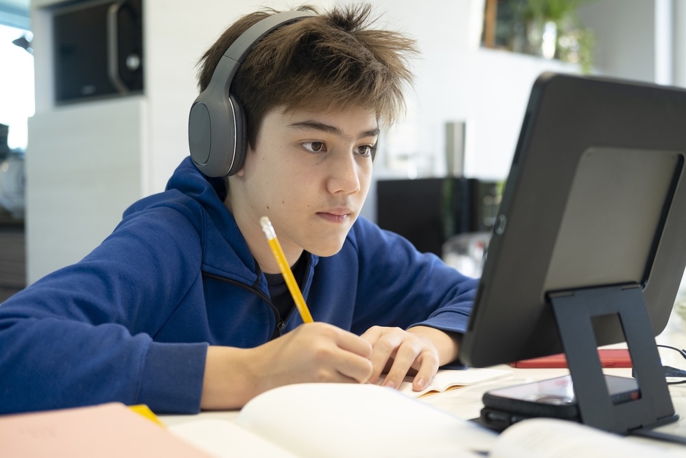 Teen boy with short brown hair wearing gray over-ear headphones and a blue hoodie, sitting at a desk and focusing intently on a tablet screen. He holds a pencil in one hand and writes in an open notebook, surrounded by books and papers in a well-lit indoor setting..