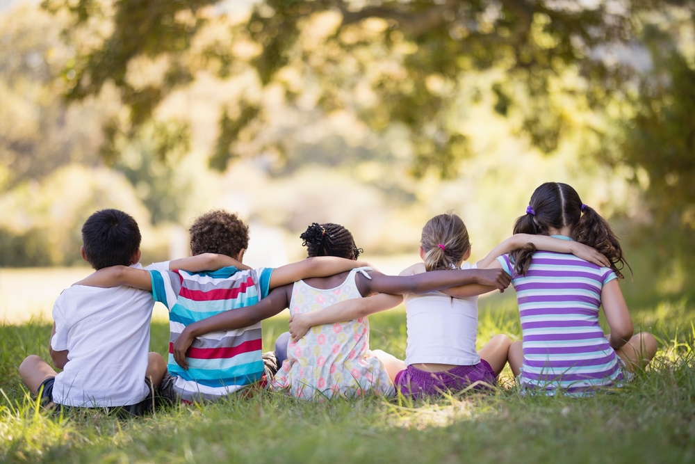 Rear view of five diverse children sitting in grass with their arms around each other under a leafy tree.