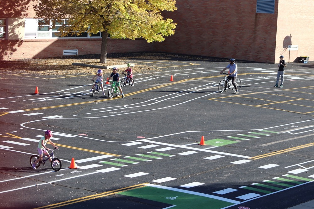 Children and adults riding bicycles through a painted practice course at the Safer Streets Practice Park outside the East Community Center. The course features miniature road markings, bike lanes, crosswalks, and traffic cones under a tree with autumn leaves.