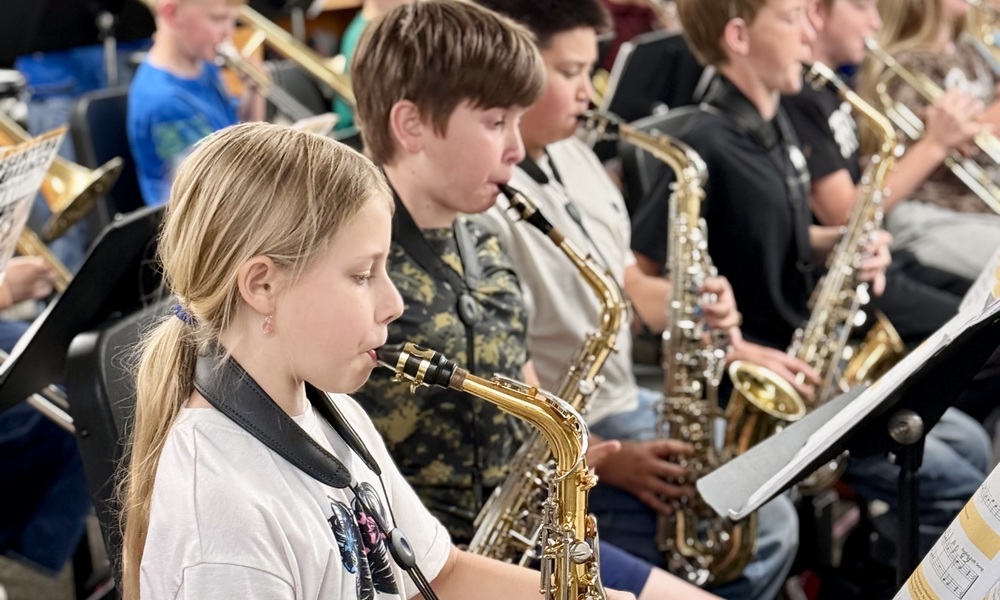 A row of middle school students playing saxophones during a band rehearsal in a music classroom.