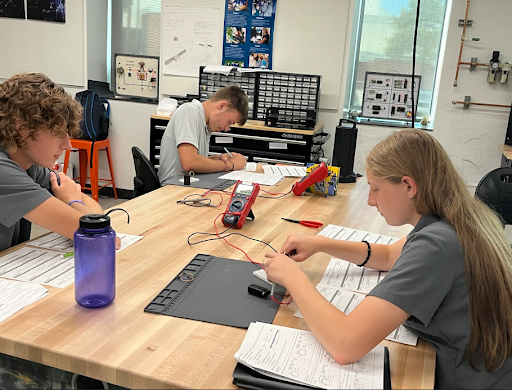 Three students are seated around a wooden table in a classroom setting, working on an electronics project. One student is using a red digital multimeter and a breadboard, another is writing on a paper, and the third is looking on.