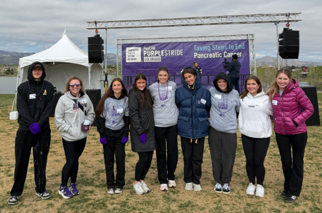 Nine people pose in front of a PanCAN PurpleStride Colorado banner and stage during a pancreatic cancer walk outdoors.